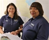 Two female City employees wearing blue shirts smiling at the camera. They both are holding paperwork. 