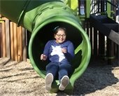 Smiling young girl wearing glasses slides through a green tube slide at the new KidsQuest playground